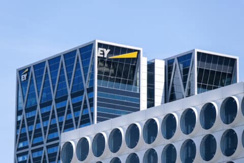Twee moderne kantoorgebouwen met glazen gevels, waaronder het in geel gemarkeerde EY-logo, staan onder een strakblauwe hemel. Het gebouw op de voorgrond heeft grote ronde ramen en is een voorbeeld van EY's toewijding aan innovaties op het gebied van energiebeheer.