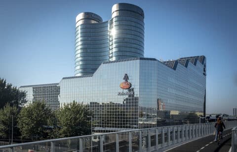 Een modern glazen kantoorgebouw met het logo van de Rabobank op de gevel, met twee cilindrische torens die boven de hoofdstructuur uitsteken. Op de voorgrond benadrukken bomen en een fietser een levendige omgeving voor de medewerkers van de Rabobank.