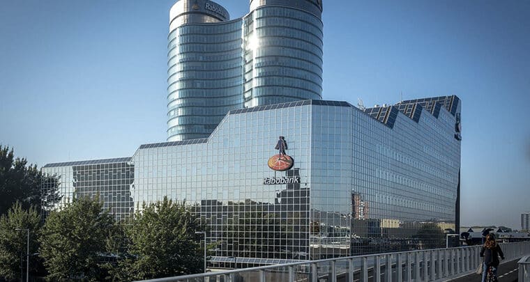 Een modern glazen kantoorgebouw met het logo van de Rabobank op de gevel, met twee cilindrische torens die boven de hoofdstructuur uitsteken. Op de voorgrond benadrukken bomen en een fietser een levendige omgeving voor de medewerkers van de Rabobank.
