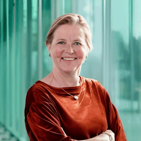 Een lachende vrouw met kort blond haar, onderdeel van het Rabobank-team, draagt een roestbruine fluwelen top en staat voor grote, groen getinte ramen, met haar armen over elkaar. De achtergrond is zacht wazig.