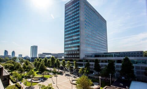 Een hoog, modern kantoorgebouw met veel ramen staat in het stadsgezicht. Het is de thuisbasis van de medewerkers van RSM. Het is omgeven door bomen, een geplaveid plein en andere gebouwen onder een zonnige, heldere hemel.