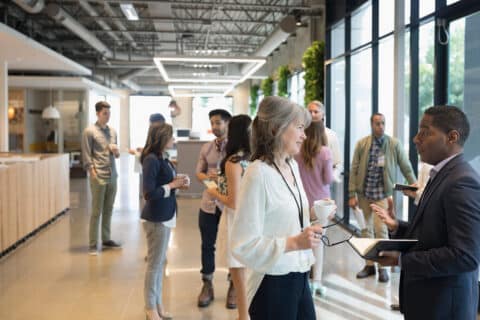 Een groep EY-professionals staat te praten in een moderne kantoorgang met grote ramen en bespreekt onderwerpen zoals energiemanagement. Sommigen houden koffiekopjes vast, anderen gebruiken apparaten in een ongedwongen, collaboratieve sfeer.