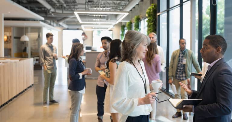 Een groep EY-professionals staat te praten in een moderne kantoorgang met grote ramen en bespreekt onderwerpen zoals energiemanagement. Sommigen houden koffiekopjes vast, anderen gebruiken apparaten in een ongedwongen, collaboratieve sfeer.