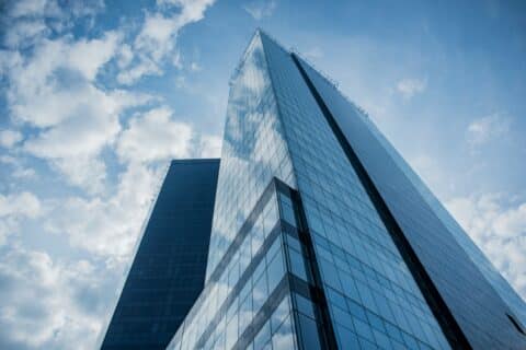 Zwei moderne gläserne Wolkenkratzer spiegeln von unten betrachtet den blauen Himmel und die weißen Wolken wider und verkörpern mit ihrem schlanken, geometrischen Erscheinungsbild vor dem Hintergrund der verstreuten Wolken das Engagement von EY für innovatives Energiemanagement.