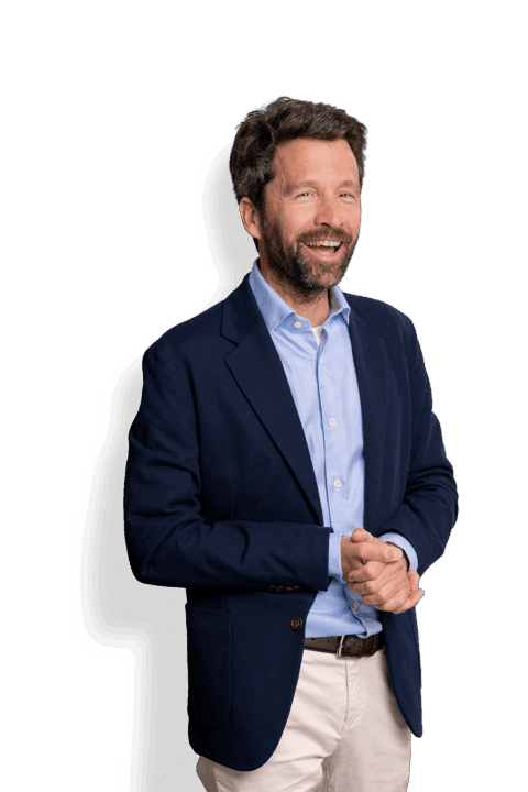 Ein Mann mit kurzen braunen Haaren und Bart steht lächelnd da. Er trägt einen marineblauen Blazer, ein hellblaues Hemd und eine beige Hose. Vor einem weißen Hintergrund hat er die Hände vor der Brust verschränkt.