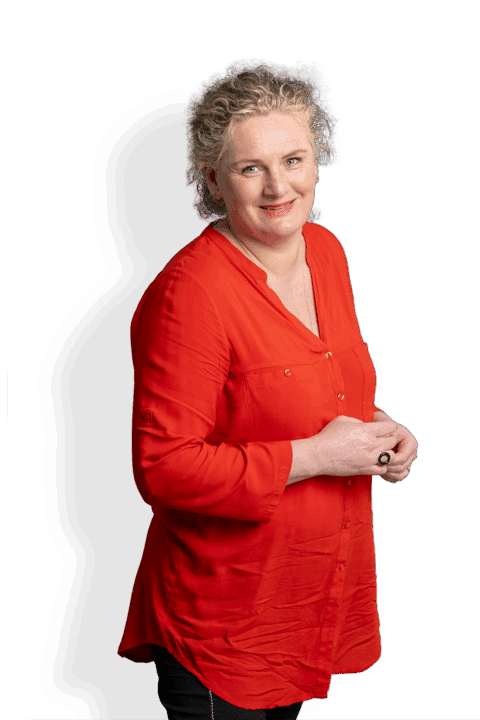Eine Frau mit lockigem blondem Haar, die eine langärmelige, leuchtend rote Bluse trägt, lächelt, während sie vor einem schlichten weißen Hintergrund steht. Sie blickt in die Kamera und hat die Hände leicht gefaltet.