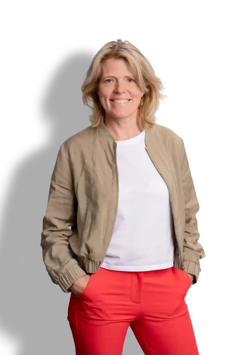 Een vrouw met blond haar glimlacht, gekleed in een lichtbruin jasje over een wit overhemd en een felrode broek, met haar handen in haar zakken, staand tegen een effen zwarte achtergrond. Dit legt de tijdloze stijl van José Janssen perfect vast.