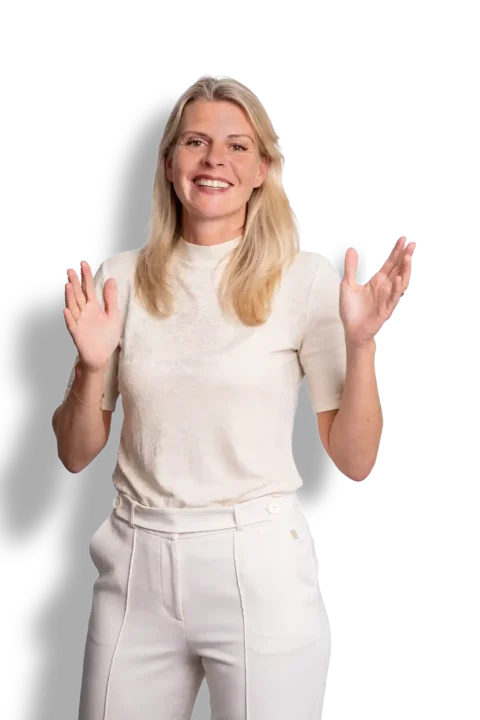 Wilma Overbeek, met lang blond haar, glimlacht en maakt gebaren met beide handen omhoog. Ze draagt een wit shirt met korte mouwen en een witte broek met hoge taille, en staat tegen een effen zwarte achtergrond.