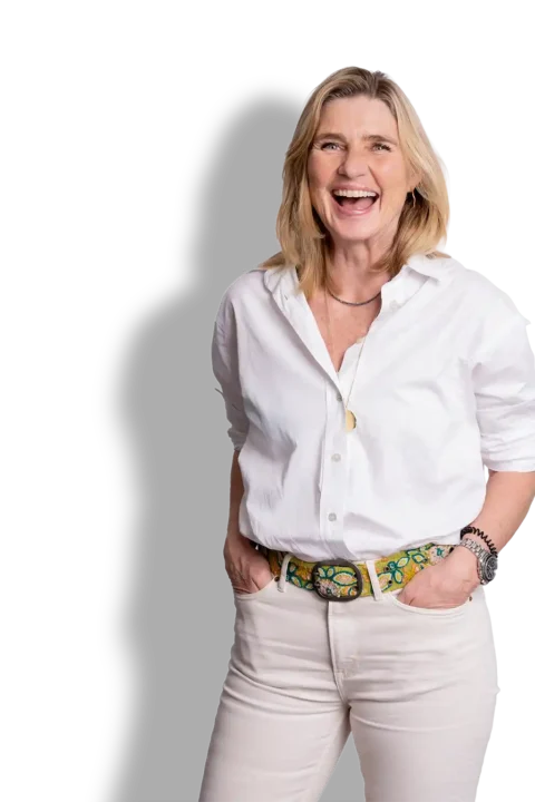 Wilma Overbeek, een vrouw met blond haar tot op haar schouders, lacht breed. Ze draagt een wit overhemd, een lichte broek en een kleurrijke riem en heeft haar handen in haar zakken.