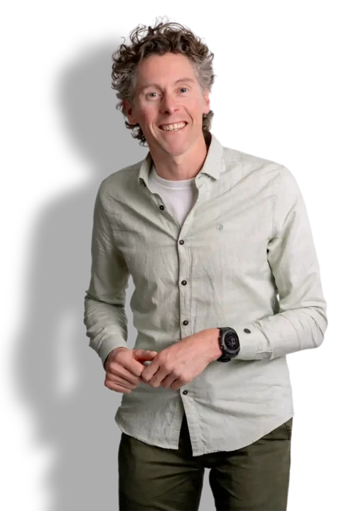 Een lachende man met krullend haar, geïdentificeerd als Erik van den Haak, draagt een lichtgroen overhemd, een wit onderhemd, een donkergroene broek en een zwart horloge. Hij staat tegen een effen zwarte achtergrond.