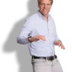 A smiling man with blonde hair, Harro Yestra, wearing a light blue button-up shirt and light gray pants, stands with his hands raised playfully, as if dancing or posing energetically.
