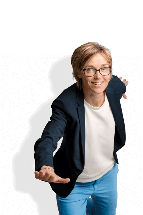 Karin Legemate, een glimlachend persoon met kort blond haar en een bril, draagt een marineblazer, wit overhemd en blauwe broek terwijl ze speels poseert met uitgestrekte armen alsof ze doet alsof ze vliegt.