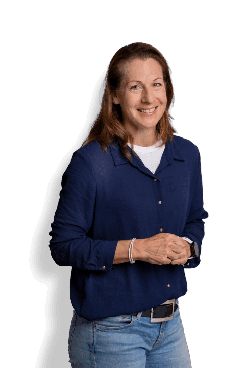 Eine Frau mit hellbraunem Haar, die ein marineblaues Hemd über einem weißen Top und blaue Jeans trägt, steht lächelnd vor einem schlichten weißen Hintergrund und hat die Hände locker vor sich gefaltet.