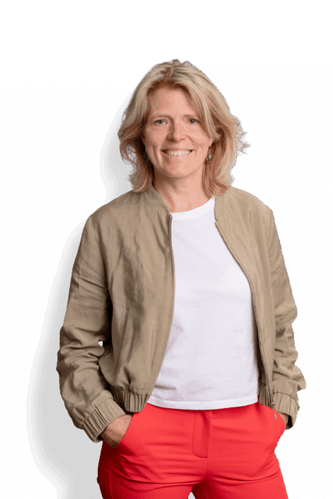 Eine Frau mit blondem Haar, die eine hellbraune Jacke über einem weißen Hemd und leuchtend roten Hosen trägt, lächelt mit den Händen in den Taschen in die Kamera und fängt den charakteristischen Stil von José Janssen ein.