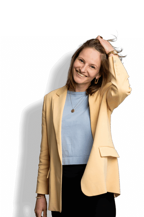 Eine lächelnde Frau, gestylt von Robin van Schaik, trägt einen gelben Blazer, ein hellblaues Oberteil und eine schwarze Hose und steht mit einer Hand vor einem schlichten Hintergrund in ihrem Haar.
