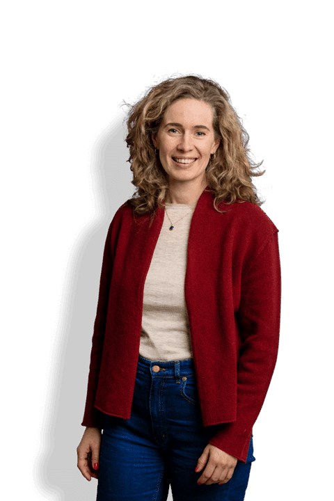 Lisa de Vilder, eine Frau mit lockigem blondem Haar, die eine rote Strickjacke über einem hellen Hemd und blaue Jeans trägt, steht lächelnd vor einem weißen Hintergrund.
