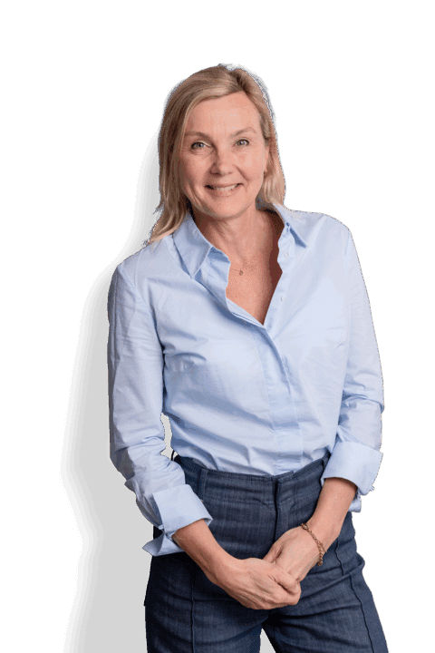Caroline Tonnaer, eine lächelnde Frau mit blondem Haar, trägt ein hellblaues Hemd mit Knöpfen und dunkle Hosen und steht mit einer Hand in der Tasche vor einem schlichten weißen Hintergrund.