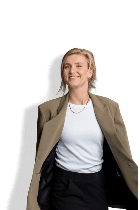 Eva van Mourik, eine lächelnde Frau mit blondem Haar, trägt einen hellbraunen Blazer über einem weißen Oberteil und einer schwarzen Hose, dazu goldene Halsketten und steht vor einem schlichten Hintergrund.