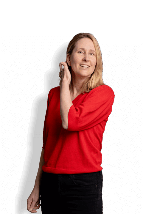 Eine lächelnde Frau mit blondem Haar, die ein leuchtend rotes Oberteil und eine schwarze Hose trägt, steht vor einem schlichten weißen Hintergrund und berührt mit einer Hand ihr Haar.