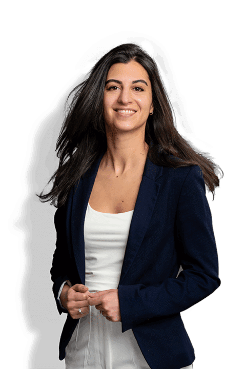 Azadeh Arooni, eine Frau mit langem, dunklem Haar, die einen marineblauen Blazer über einem weißen Oberteil trägt, steht selbstbewusst lächelnd da und hält mit den Händen locker ihre Jacke.