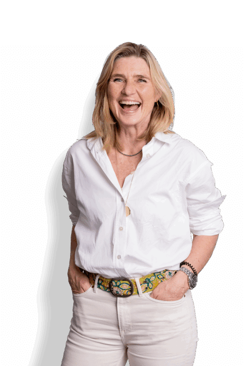 Wilma Overbeek, een lachende vrouw met blond haar, draagt een wit overhemd, een riem met print en een lichte broek. Ze staat met haar handen in haar zakken tegen een witte achtergrond.