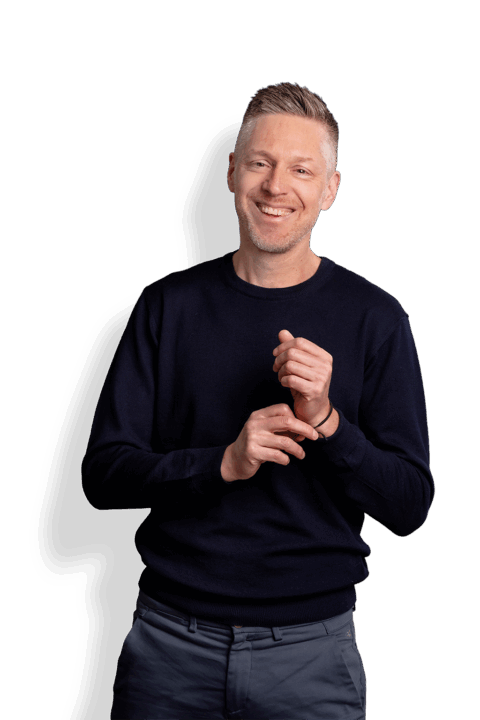 Ein lächelnder Mann mit kurzen, hellen Haaren, einem marineblauen Pullover und grauen Hosen steht vor einem weißen Hintergrund und hält mit der rechten Hand sein linkes Handgelenk.