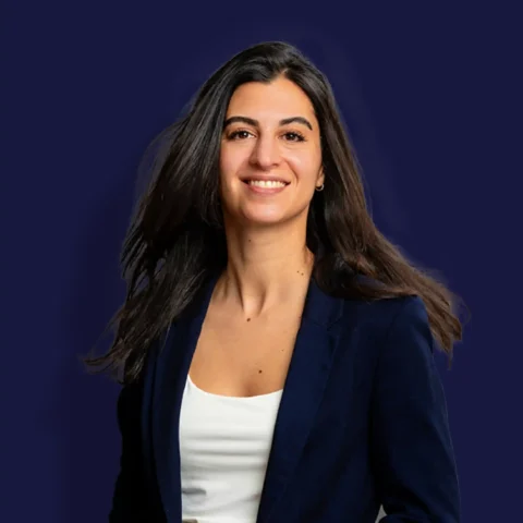 Een vrouw met lang donker haar, gekleed in een marineblauwe blazer en een wit topje, lacht zelfverzekerd tegen een effen donkerblauwe achtergrond.
