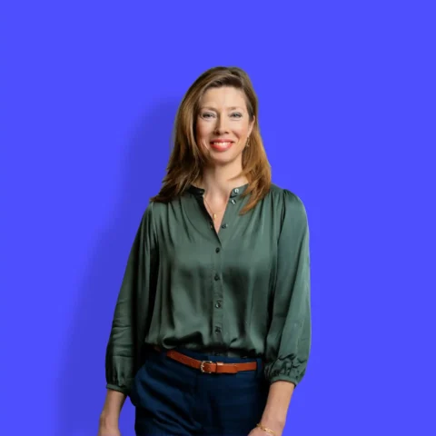 Een vrouw met lichtbruin haar, gekleed in een donkergroene blouse en een zwarte broek, staat lachend voor een effen, helderblauwe achtergrond.