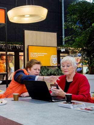 Twee vrouwen zitten aan een tafel met een laptop en koffiekopjes. Een van hen, in het oranje, wijst naar de telefoon van de vrouw in het rood, mogelijk pratend over het ING Lifeguardprogramma. De lichte, binnenomgeving is voorzien van digitale schermen op de achtergrond.