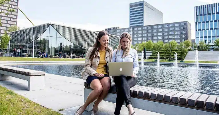 Twee vrouwen, mogelijk medewerkers van RSM, zitten op een bankje bij een vijver met fonteinen en kijken naar een laptop. Moderne kantoorgebouwen en bomen zijn op de achtergrond zichtbaar onder een heldere hemel.