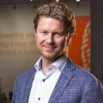 Een man met golvend blond haar en een korte baard, gekleed in een blauw geblokte blazer en een wit overhemd, staat binnen met op de achtergrond wazig oranje en witte muurreclame, die doet denken aan een stijlvolle badmeester die buiten dienst is.
