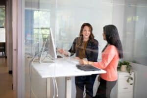 Zwei Frauen stehen in einem modernen Büro an einem Schreibtisch mit Computer und unterhalten sich. Der Blick ist durch eine Glaswand zu sehen. Eine Frau lächelt, während die andere zuhört. Auf dem Schreibtisch sind Papiere und Pflanzen zu sehen.