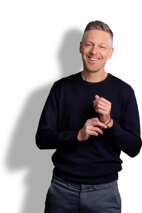 Een glimlachende man met kort haar, gekleed in een donkere trui en een grijze broek, staat tegen een zwarte achtergrond en trekt met één hand zijn mouw recht.