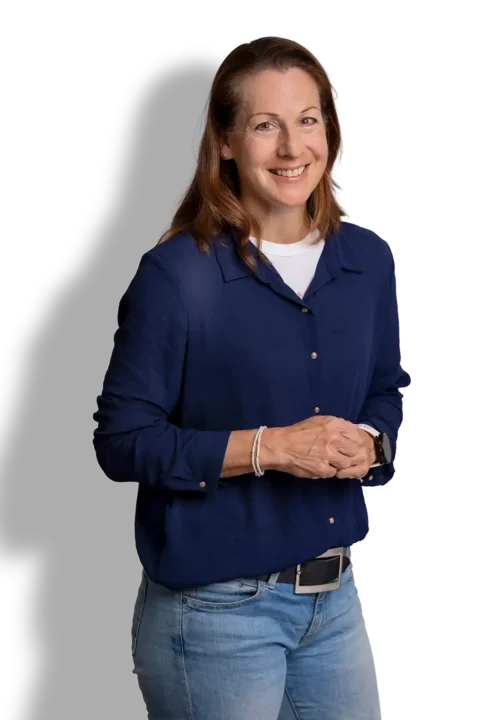 Een vrouw met lichtbruin haar, gekleed in een marineblauw overhemd over een wit topje en een lichte spijkerbroek, staat lachend met haar handen losjes gevouwen voor zich, tegen een effen achtergrond.