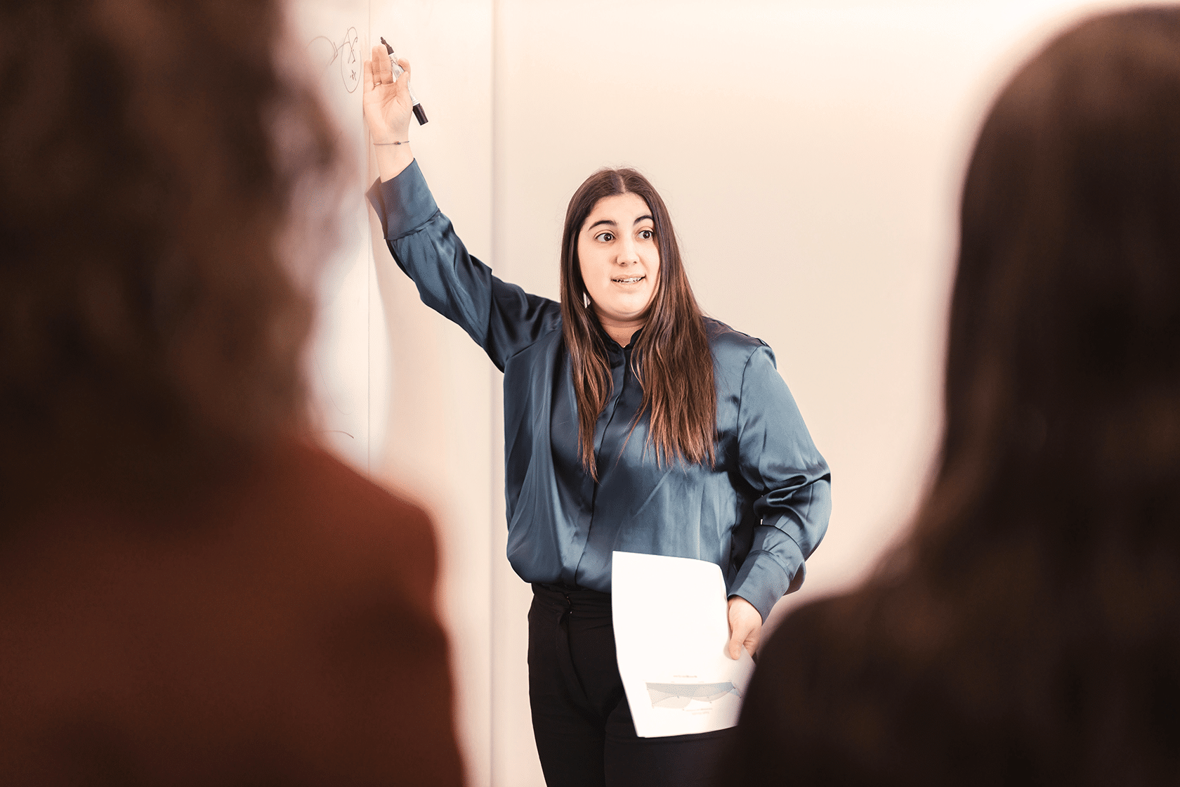 Een vrouw in een blauwe blouse staat bij een whiteboard, houdt papieren en een marker vast terwijl ze twee mensen toespreekt in een klaslokaal of vergaderomgeving van een Top Mom programma. Ze gebaart naar het bord terwijl ze iets uitlegt.