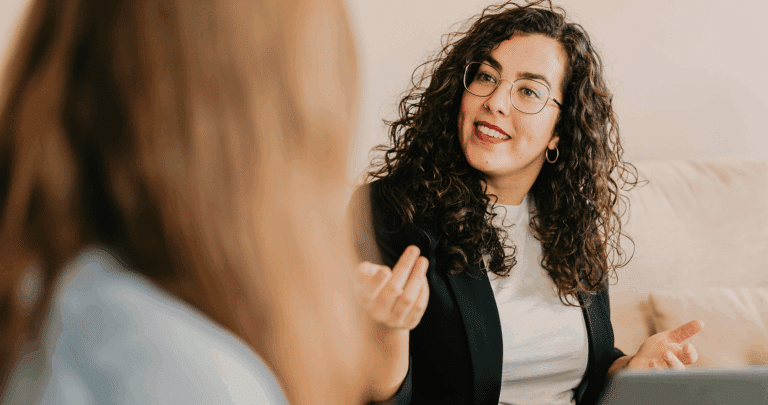 Een vrouw met krullend haar en een bril, met een zwarte blazer en wit overhemd, zit op een bank en maakt gebaren terwijl ze het Top Mom programma bespreekt met een andere persoon die met haar rug naar de camera staat.