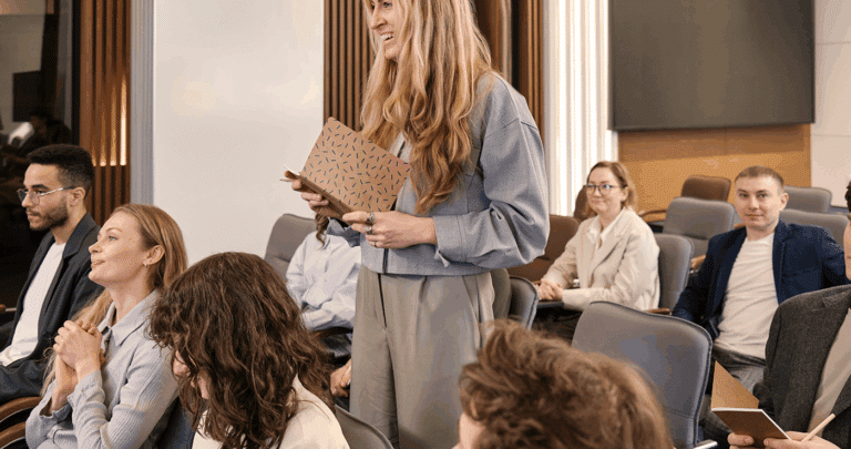 Een vrouw staat en glimlacht terwijl ze een notitieblok vasthoudt in een moderne vergaderzaal vol zittende mensen die bezig zijn met het Top Mom programma en deelnemen aan een aandachtige discussie.