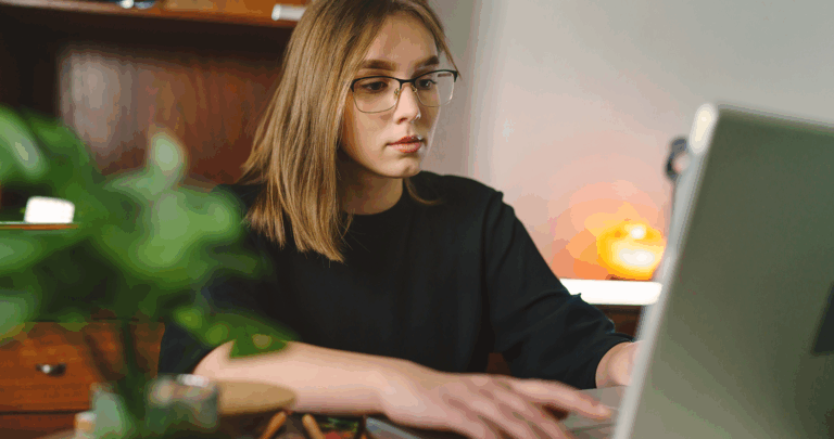 Een jonge vrouw met bril zit aan een bureau, geconcentreerd op een laptop terwijl ze aan haar programma werkt. Ze typt met beide handen. De achtergrond bestaat uit planken, een lamp en een groene potplant op de voorgrond.