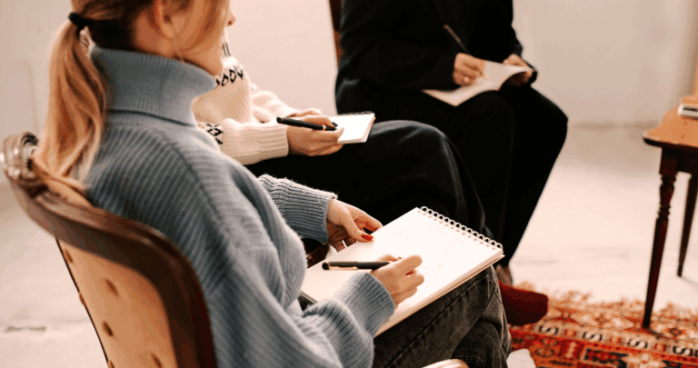 Drie mensen zitten in een stoel, met notitieboekjes en pennen in de hand, bezig met schrijven of aantekeningen maken - misschien tijdens een Top Mom-sessie. De focus ligt op hun handen en schoot; warme truien en een tapijt met patroon maken de sfeer nog gezelliger.
