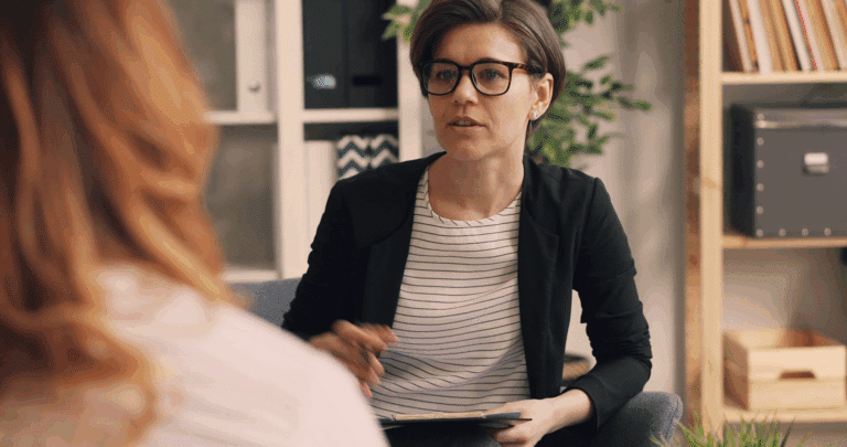 Een vrouw met kort haar en een bril, gekleed in een blazer en gestreept overhemd, zit binnen met een klembord en pen, aandachtig luisterend naar een andere persoon tijdens een Top Mom programmasessie.