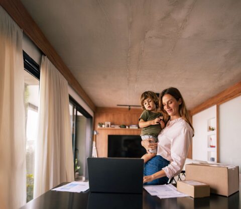 Een vrouw staat achter een bureau met een jong kind in de hand en kijkt naar een laptop. Er liggen papieren en een kartonnen doos op het bureau terwijl het zonlicht door de grote ramen schijnt - een perfecte weergave van het drukke leven van een topmoeder in een modern huis.