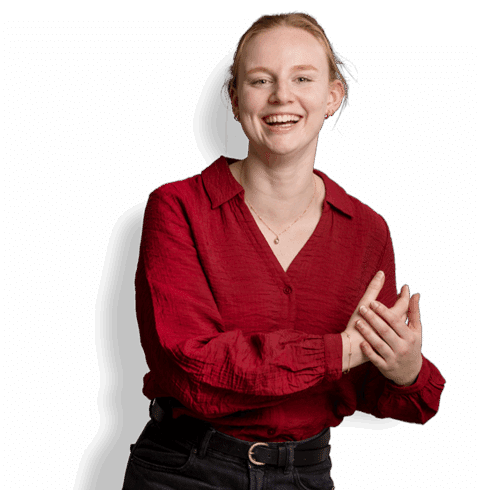 Een jonge vrouw met een lichte huid en blond haar, in een rode blouse met structuur en een zwarte broek, staat te glimlachen en klapt in haar handen tegen een effen witte achtergrond. Ze belichaamt het vertrouwen van een programma-adviseur voor deze vacature.